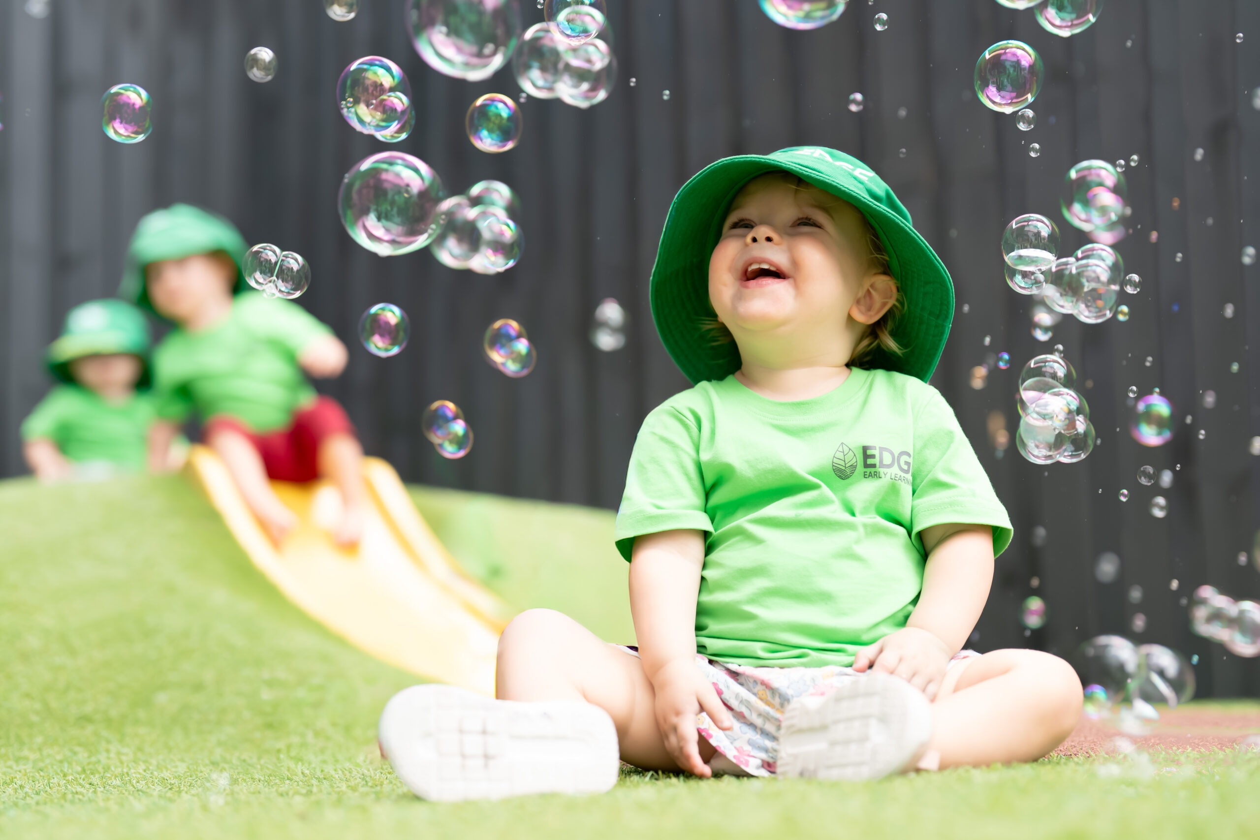 A young child in a green hat and shirt joyfully plays with bubbles at Edge Grange daycare.
