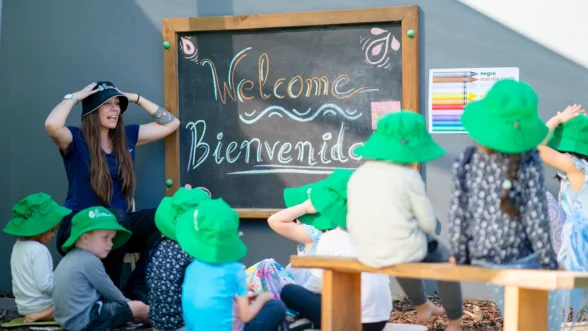 Kindergarten teacher and children in front of blackboard with the words "Welcome, Bienvenido" at Edge West End - Montague Road.
